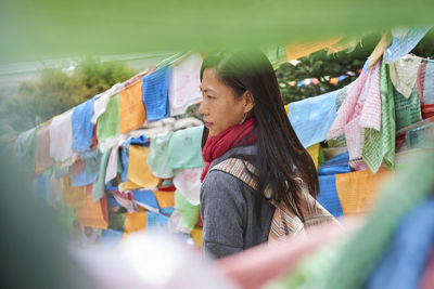 Young ethnic female tourist in warm clothing and hat looking away while crossing simple long narrow suspension wooden bridge decorated with bright multi colored pieces of fabric in daylight