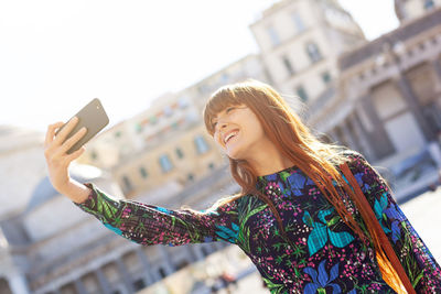 Smiling young woman taking selfie while standing against building