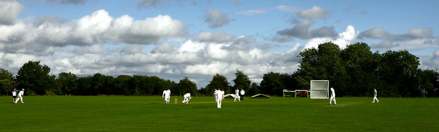 Panoramic view of people playing cricket on field against sky