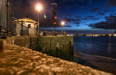 Illuminated street by sea against sky at night