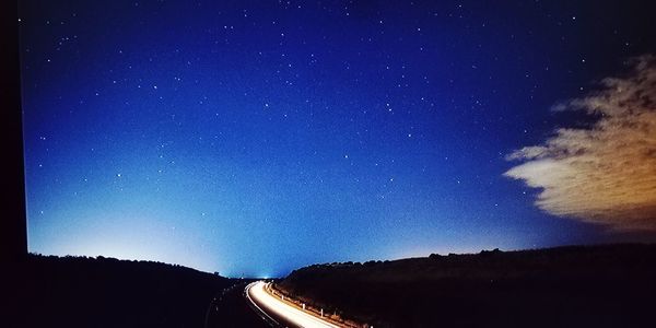 Road amidst silhouette trees against blue sky at night