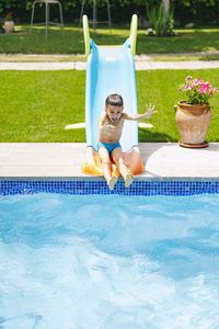 Full body of excited shirtless little boy with wet dark hair riding on water slide while spending sunny summer day in aqua park