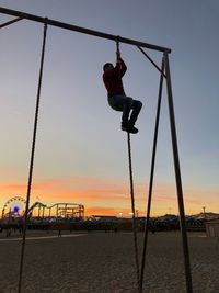Man climbing on rope at playground against sky during sunset
