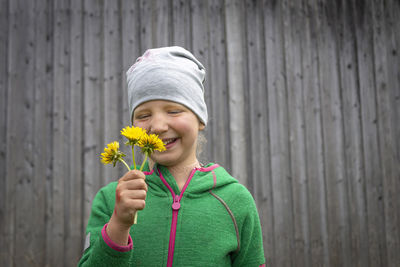 Portrait of smiling woman holding flower while standing against wooden wall