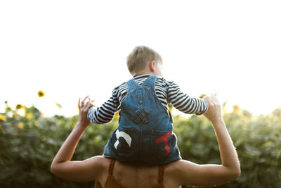 Back view of a son sitting on mother's shoulders amidst sunflower