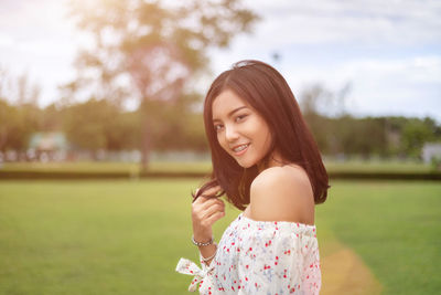 Portrait of smiling young woman standing on grass against trees and sky