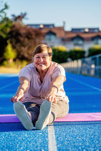 Happy smiling senior plus size woman with earphones doing stretches sitting on yoga mat outdoors 