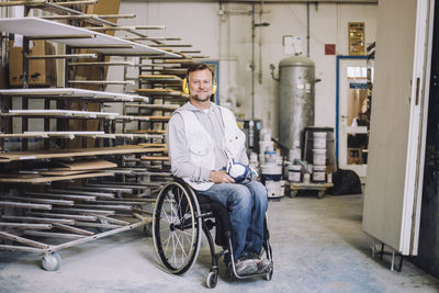 Full length of smiling painter wearing ear protectors while sitting on wheelchair in workshop