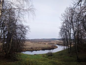 Bare trees on field by lake against sky