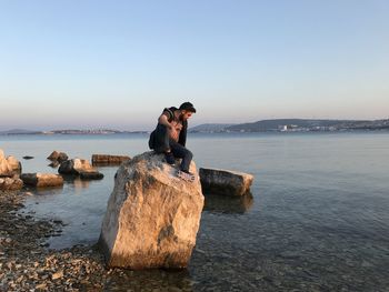 Man sitting on rock by sea against clear sky