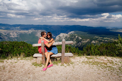 Young couple sitting on bench against mountains