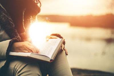Midsection of woman reading book in sea against sky during sunset
