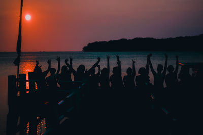 Silhouette people with arms raised enjoying at beach against sky during sunset