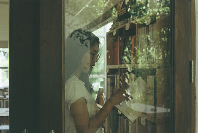 Woman taking book from bookshelf seen through window