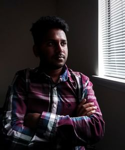 Portrait of young man looking away while standing against window