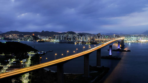 Illuminated bridge over river against cloudy sky at night