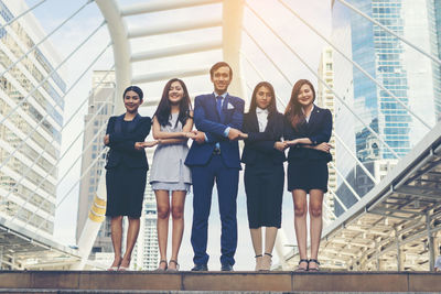 Portrait of smiling young colleagues with arms crossed holding hands on footpath against buildings in city