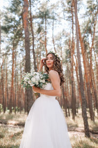 Portrait of young woman in wedding dress holding flower bouquet while standing against trees