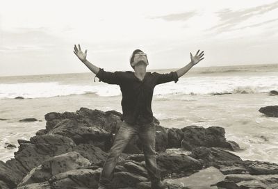 Young man standing with arms outstretched on rock formations at seashore