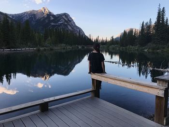 Rear view of teenage girl sitting on railing by lake against mountains