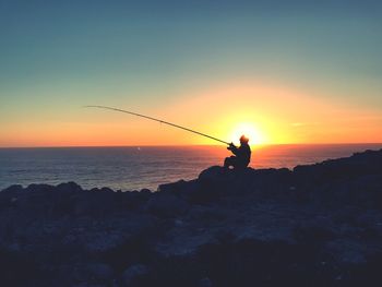 Person fishing while resting on rocky shore against clear sky during sunset