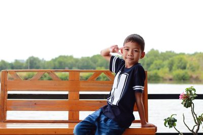 Portrait of boy gesturing peace sign while sitting on bench by lake
