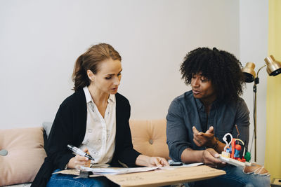 Multi-ethnic business colleagues discussing over placard while sitting on sofa in lobby at creative office