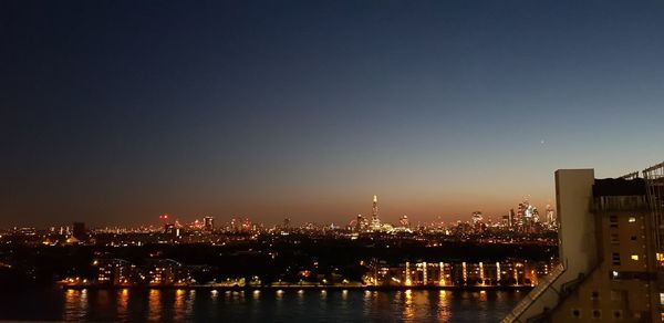 Illuminated buildings by river against clear sky at night