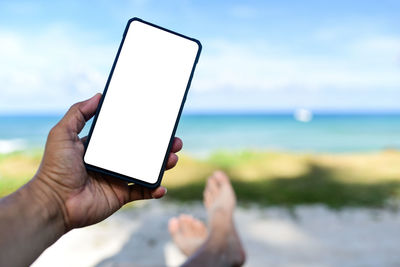 Person holding mobile phone by sea against sky