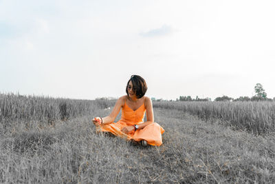 Young woman sitting on land in field against sky