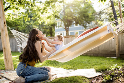 Happy mother playing with daughter sitting in hammock at yard