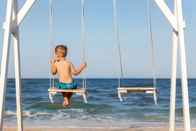 Back view  little lonely sad kid boy swinging at sea sand beach against blue ocean water clear sky.