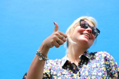 Low angle view of smiling young woman gesturing thumbs up against blue sky