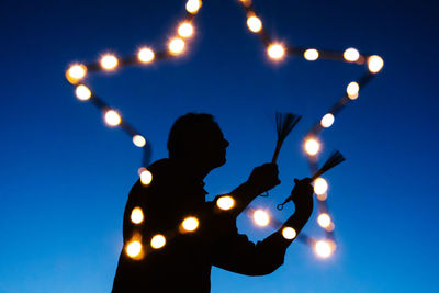 Silhouette man playing instrument seen through illuminated star shape decoration against clear blue sky at night