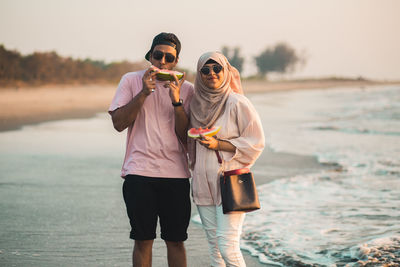 A heterosexual couple standing together on the beach and eating fruit