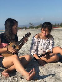 Young woman playing ukulele by friend holding mobile phone while sitting at beach