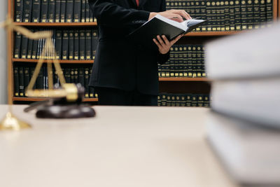 Midsection of lawyer reading book while standing against bookshelf in office