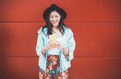 Happy smiling young woman using phone against red wall