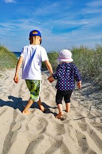 Rear view of siblings walking at beach against blue sky during sunny day