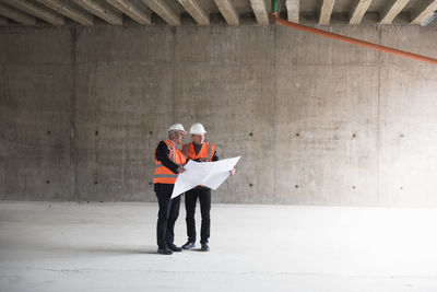 Two men with plan wearing safety vests talking in building under construction