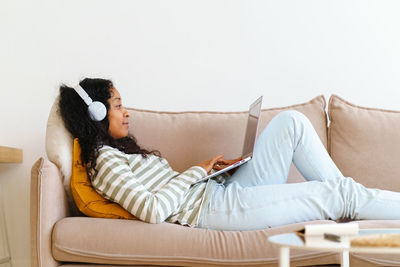 African-american female in headphones surfing the net on laptop while lying on sofa in living room