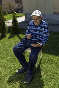 Senior man with magnifying glass and mobile phone sitting by walking cane at backyard against house