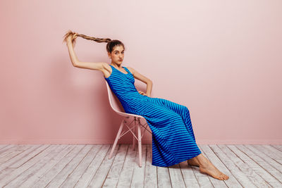 Side view of young woman in blue dress barefoot looking at camera holding ponytail while sitting with in chair and looking at camera on minimalist pink background