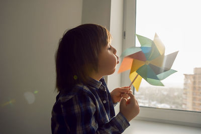 Child boy blows on a multi-colored turntable while sitting at home on the background of a window