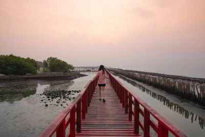 Rear view of woman walking on footbridge by lake against sky during sunset