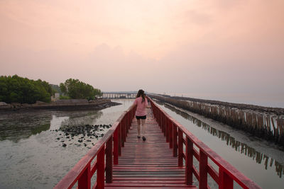 Rear view of woman walking on footbridge against sky during sunset