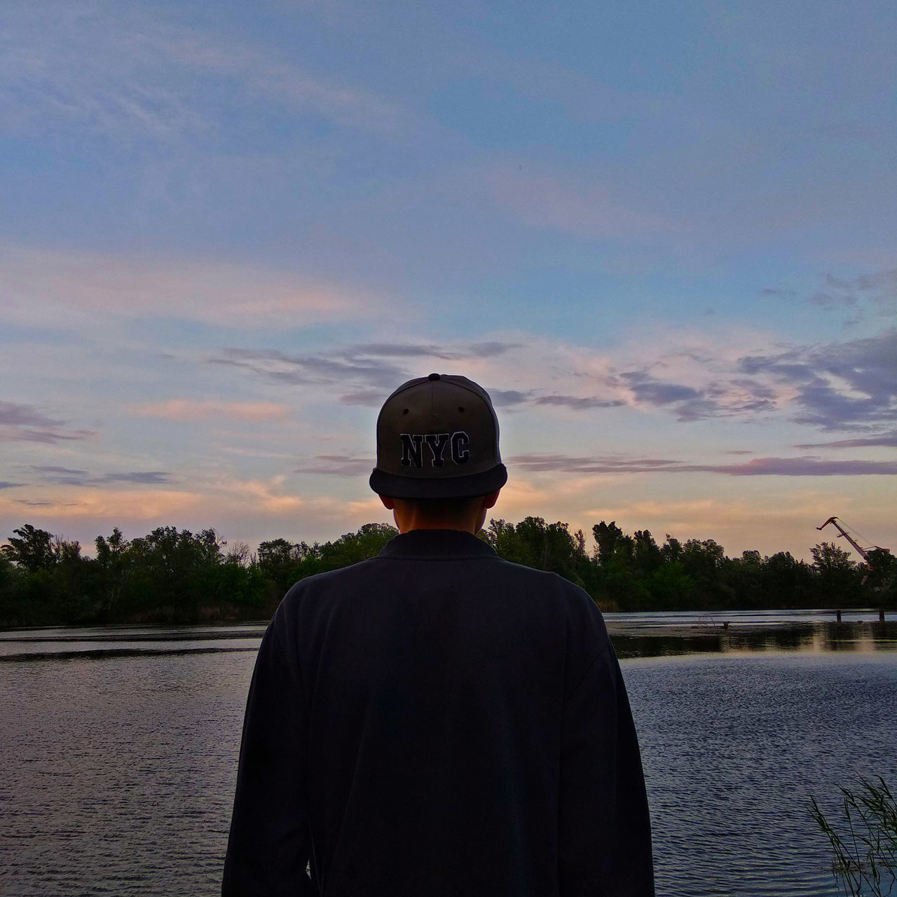 REAR VIEW OF MAN STANDING BY LAKE AGAINST SKY AT SUNSET