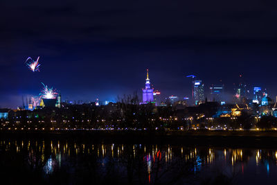 Illuminated buildings by river against sky at night