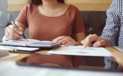 Midsection of businesswoman writing in book while colleague pointing at graph