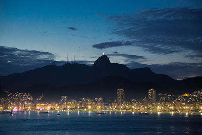 Illuminated buildings by sea against sky at dusk
